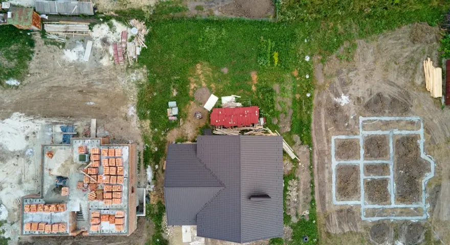 Widok z lotu ptaka na plac budowy z fundamentami, częściowo zbudowanym domem i materiałami budowlanymi.