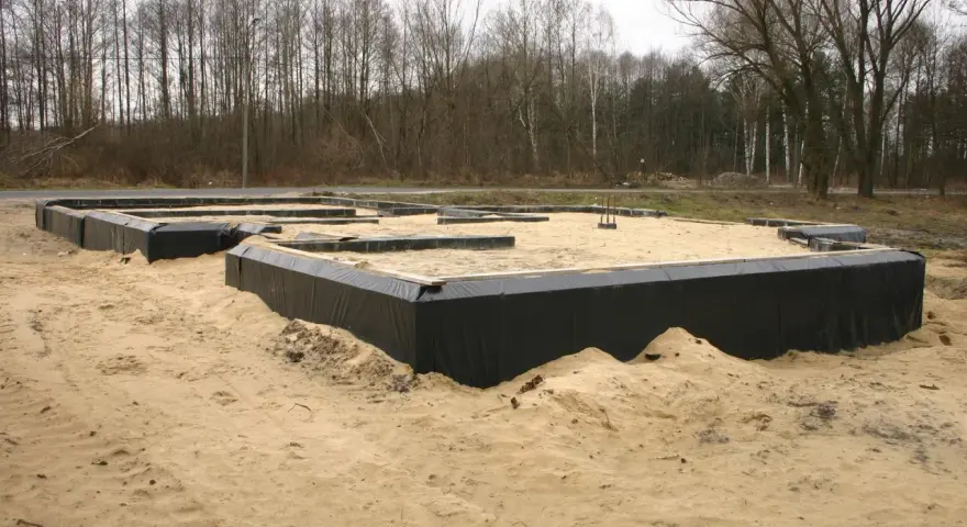 Fundamenty domu zabezpieczone czarną folią przed wilgocią. Piasek wokół budowy.
