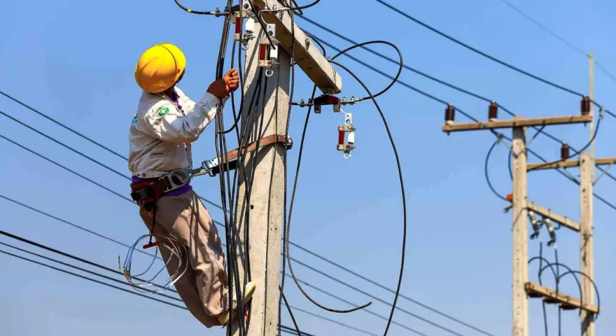Elektryk w żółtym kasku pracuje na słupie energetycznym, podłączając przewody. Zastanawiasz się, ile kosztuje przyłącze elektryczne?