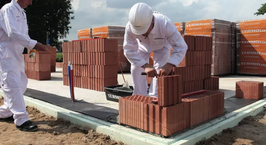 Dwóch budowlańców w białych kombinezonach układa cegły na fundamencie.